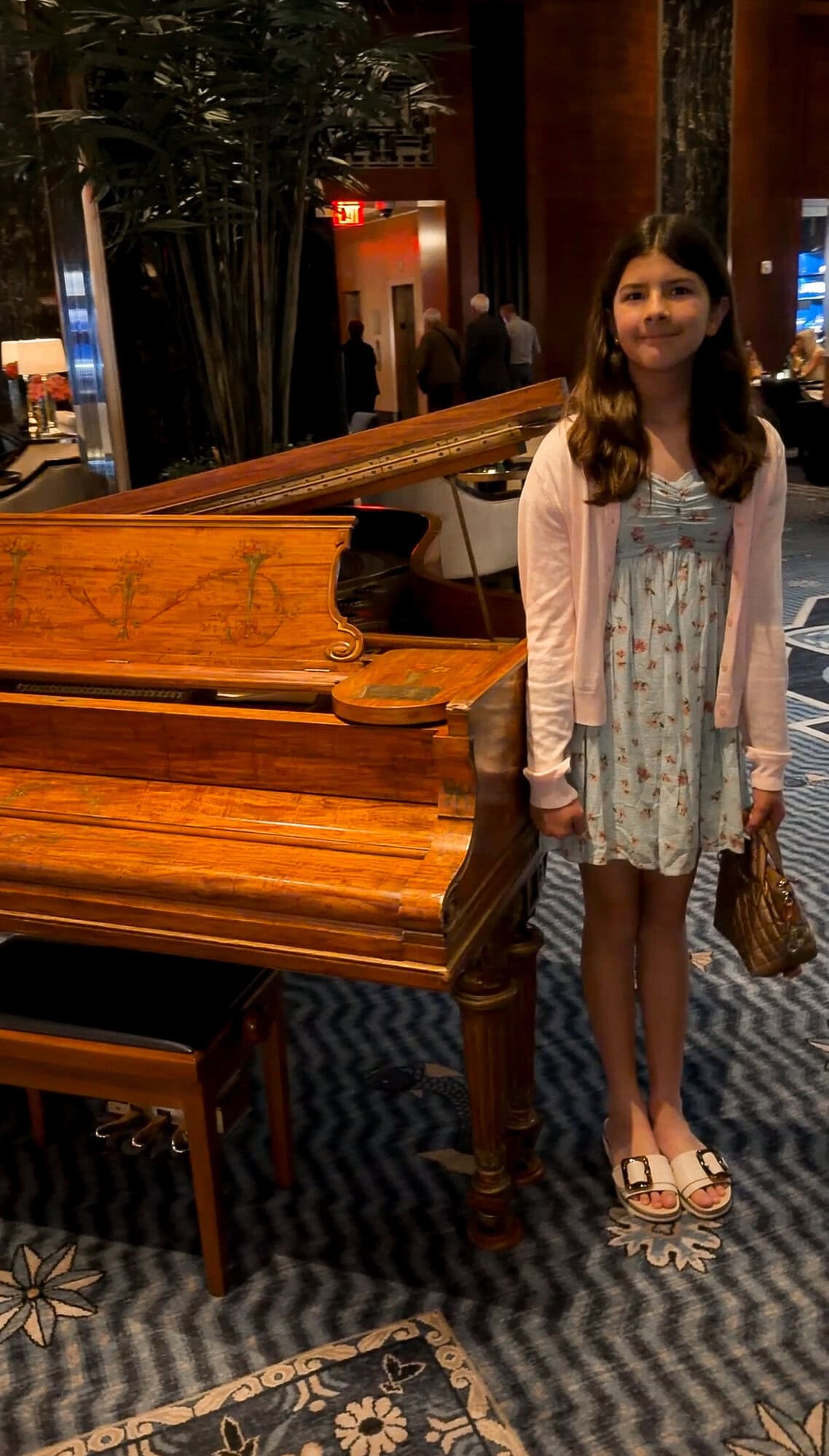 Tween daughter in a blue floral sundress and cream cardigan standing next to a grand piano in the Waldorf Astoria hotel