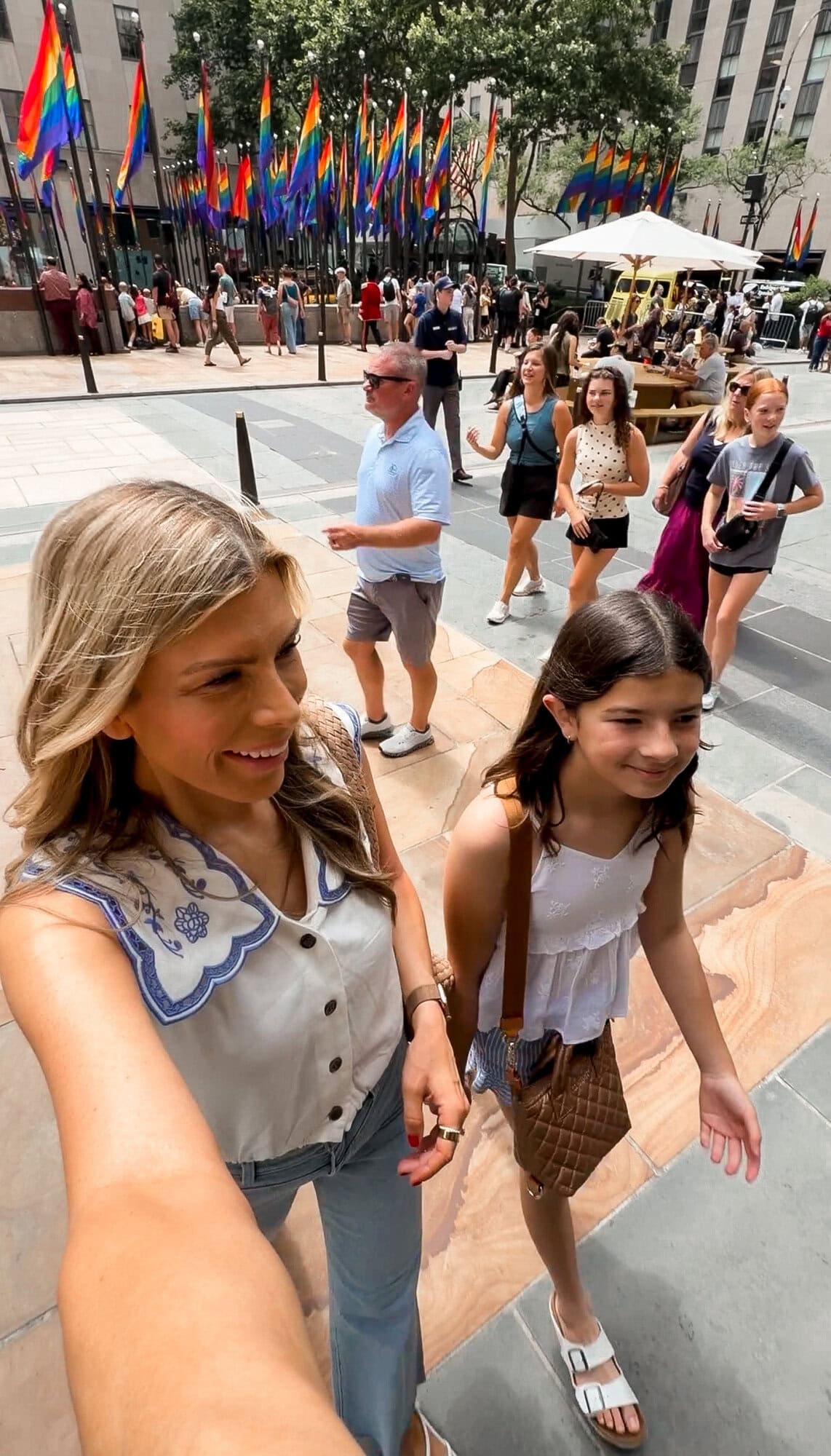 Mom and tween walking through Rockefeller Plaza with colorful flags overhead in NYC, both wearing white blouses and jeans